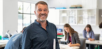 : male teacher with papers in his hand, with students working at tables in the background