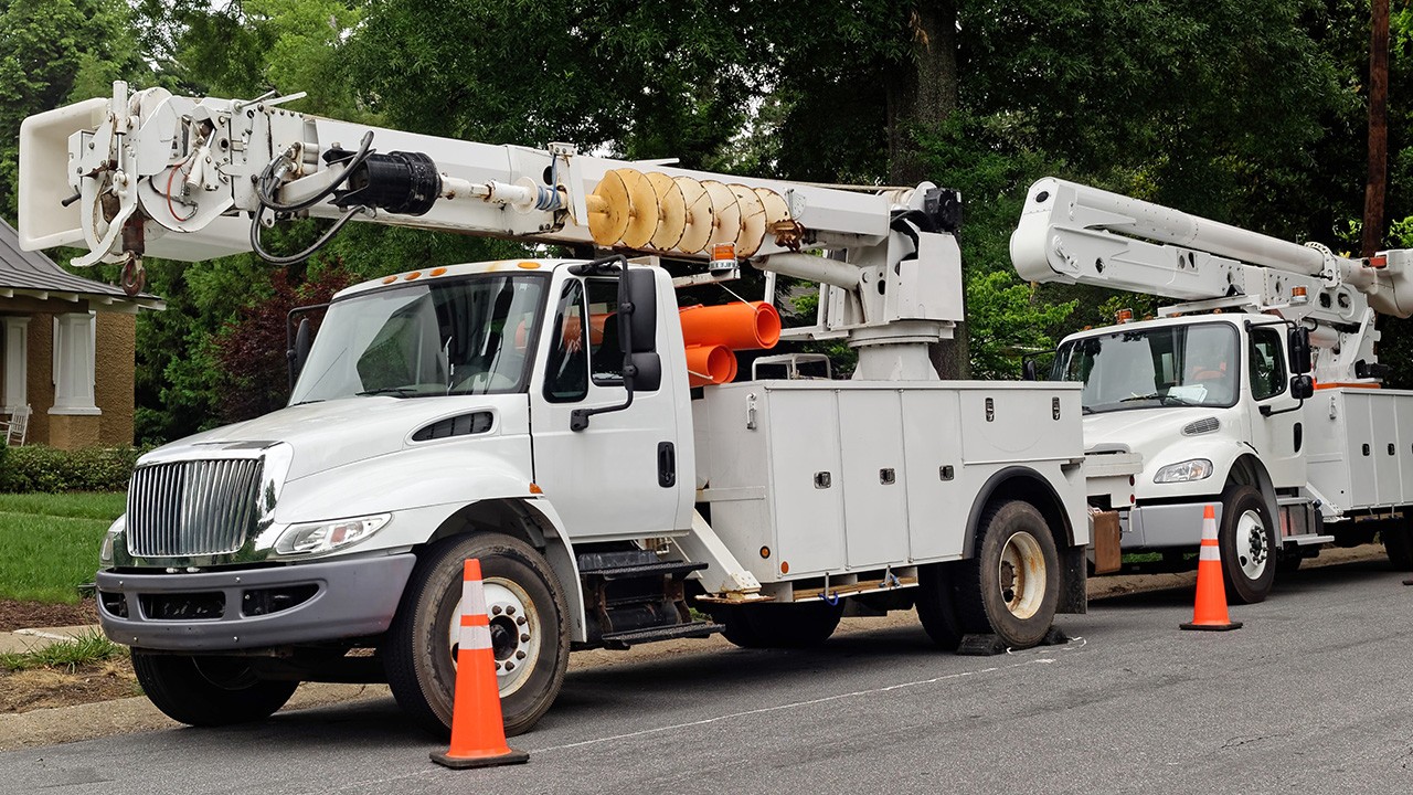 Two white trucks with buckets for aerial line installation and maintenance are parked on the road, with orange security cones around them