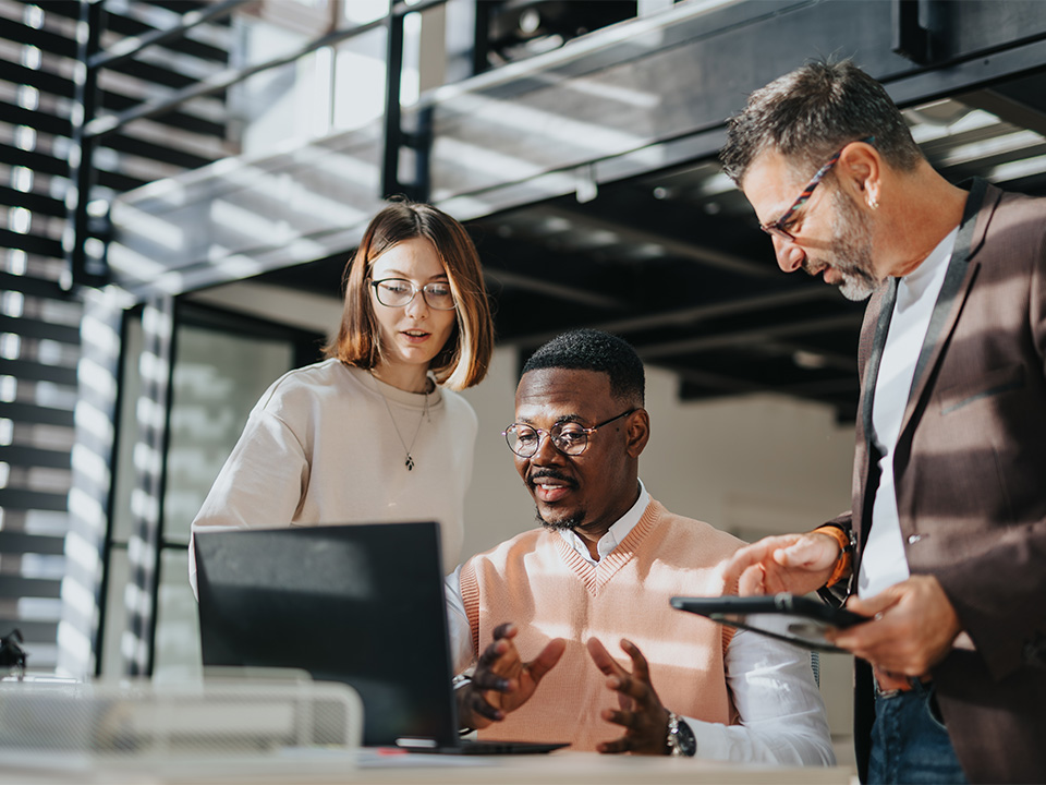 A woman and two men gather around a computer screen with sunlight filtering into the space through window blinds